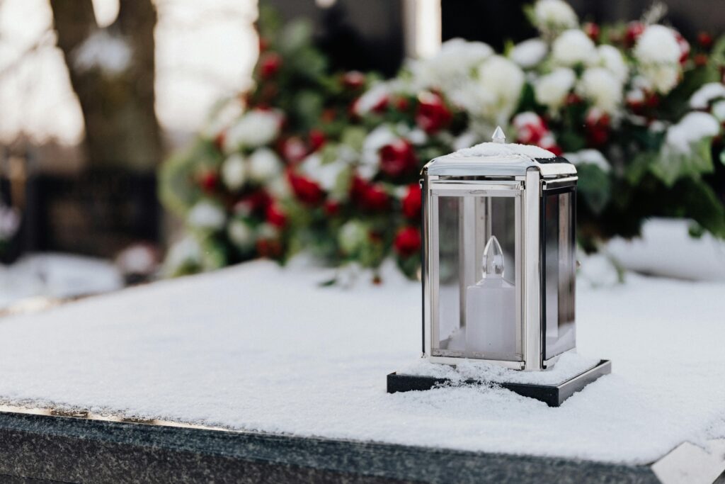 pexels-photo-6769874-6769874 A lantern in snow on a tomb, with blurred colorful flowers in the background.