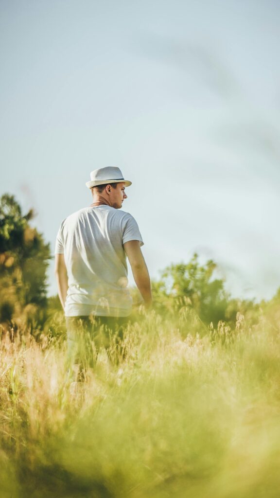 pexels-photo-11209961-11209961 A man wearing a hat stands in a sunlit field, gazing into the distance, embracing nature's tranquility.